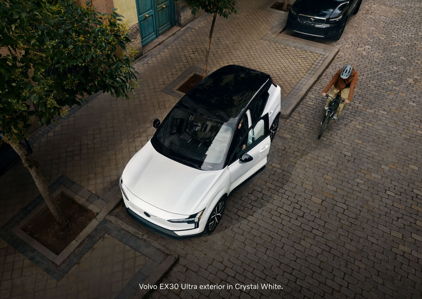 High-angle view of a white Volvo EX30 parked on a cobblestone street as a cyclist rides past.