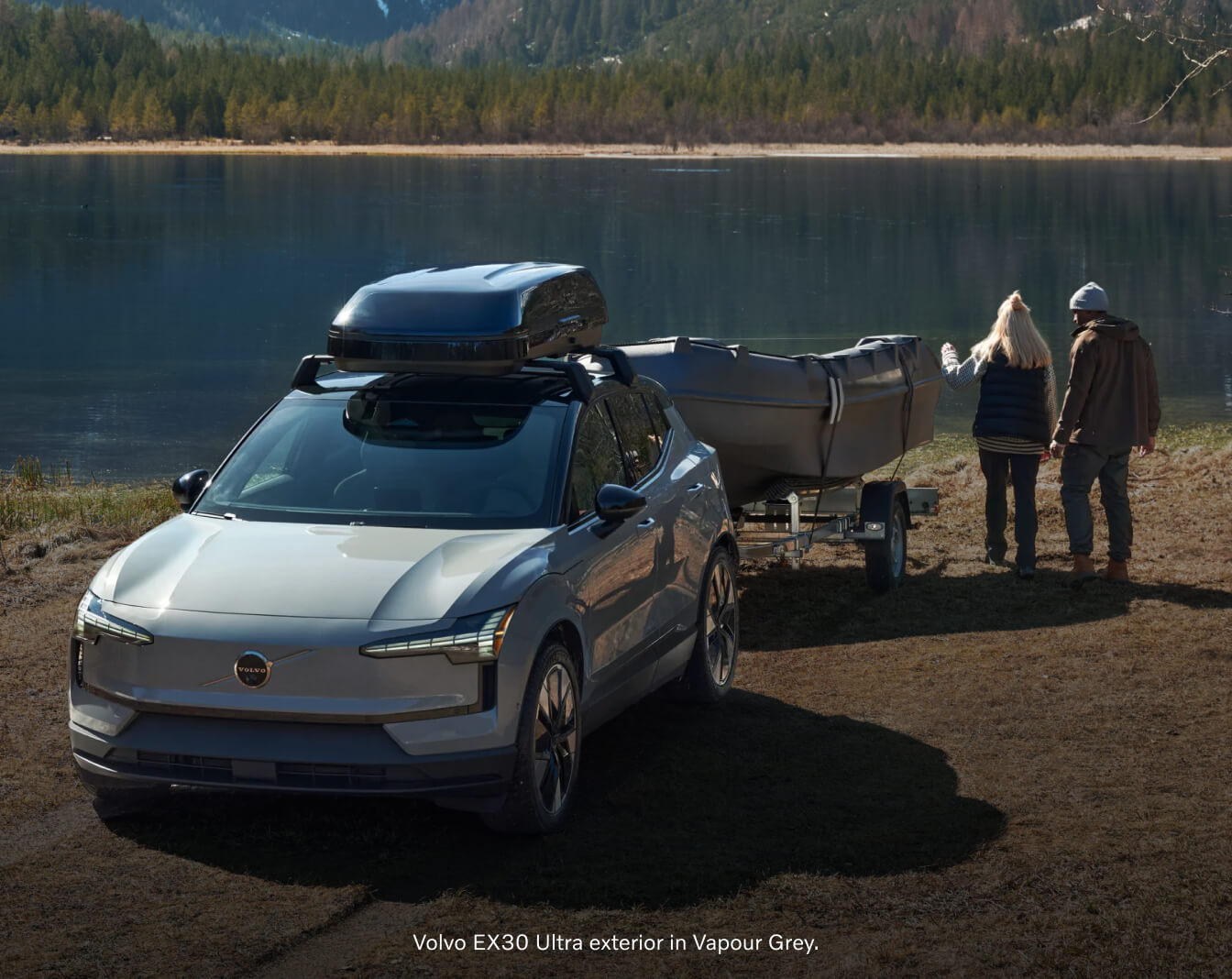 Front three-quarter view of a Volvo EX30 towing a boat trailer on a lakeside shore with a couple.