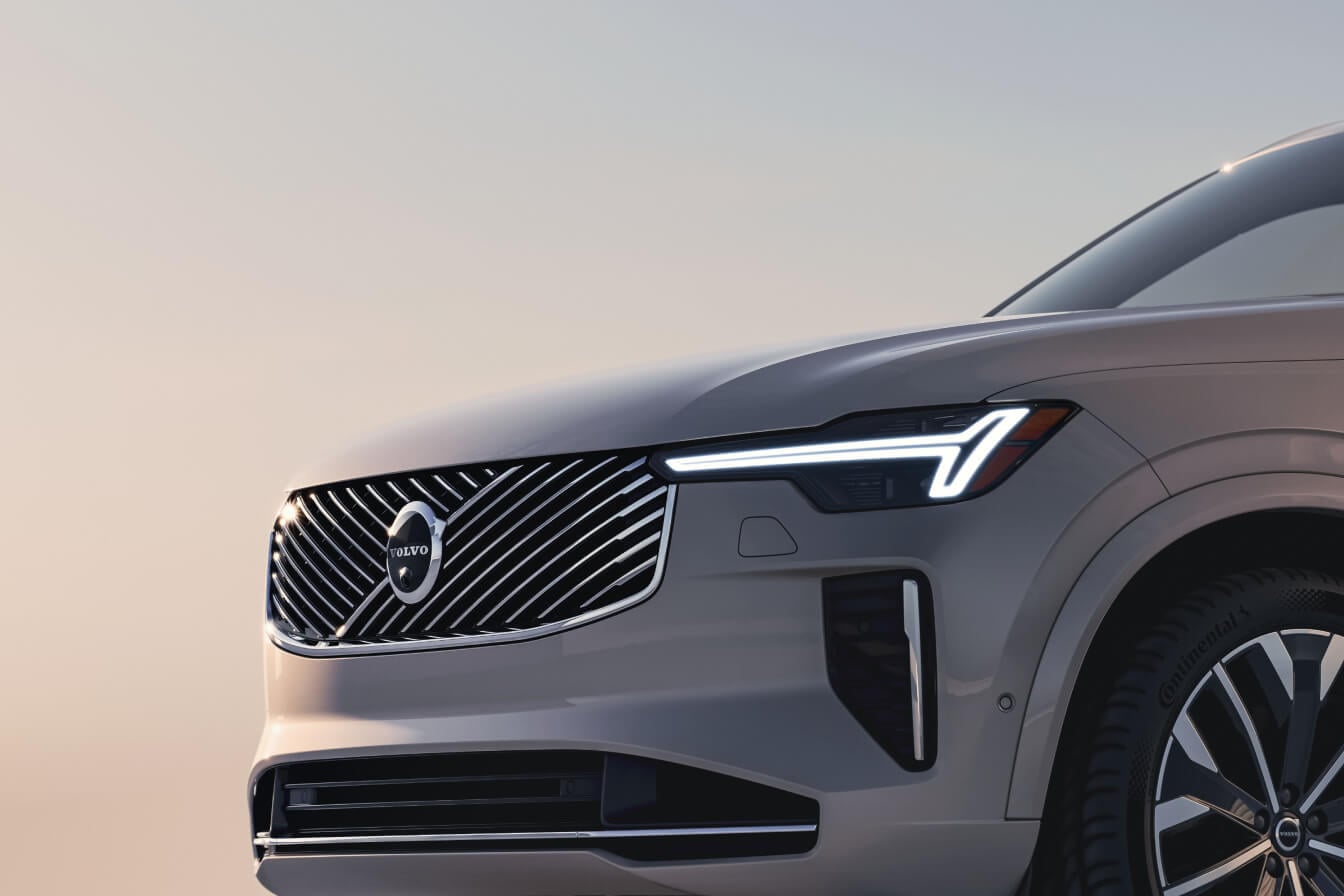 Close-up front view of a beige Volvo XC90 SUV headlight and grille against a soft sky.