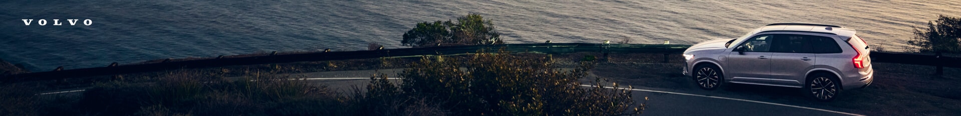 High-angle side view of a silver Volvo XC90 SUV on a coastal road overlooking the ocean at sunset.