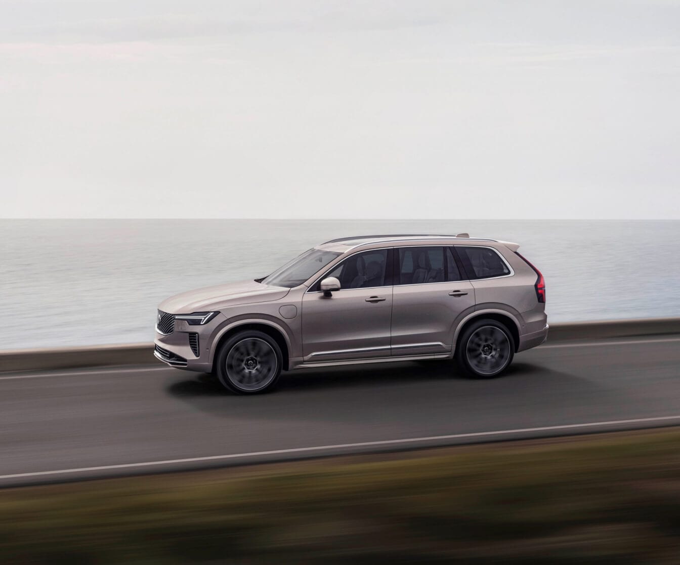 Side view of a beige Volvo XC90 SUV driving on a coastal road against an ocean background.