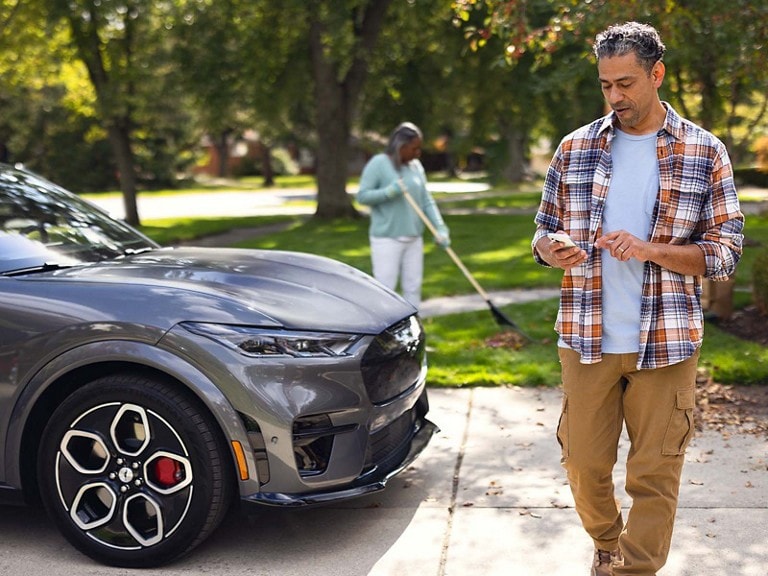 Two adults are outside near their Mustang Mach-E. One rakes leaves, while the other checks their phone