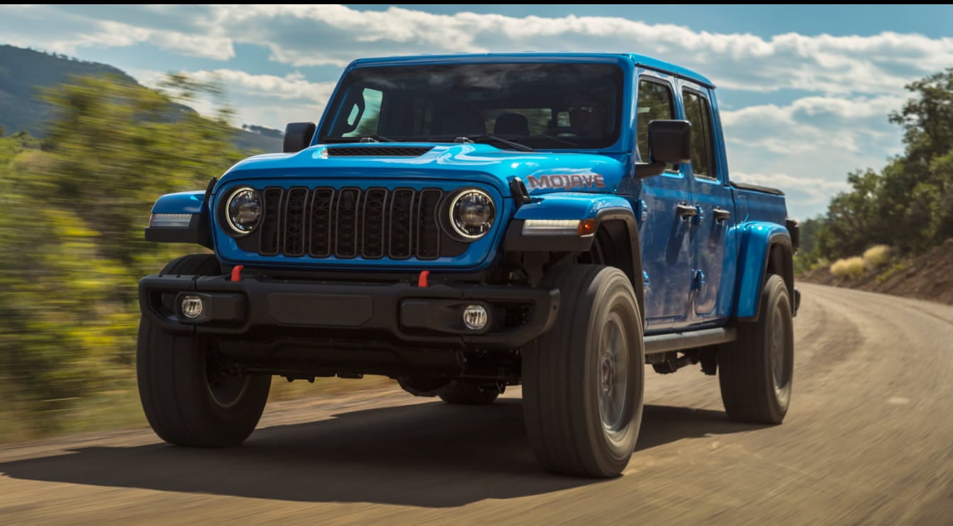 2026 Jeep Gladiator drives on a dirt road under a clear sky, surrounded by greenery. The vehicle conveys power and adventure.