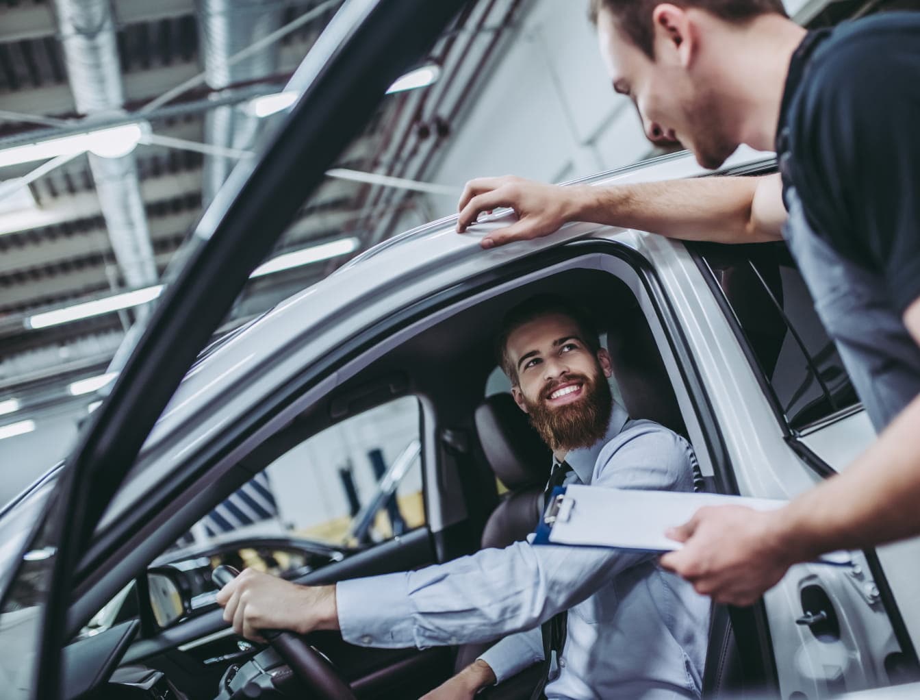 Man smiling while sitting in a car, talking to another man holding a clipboard in a garage setting.