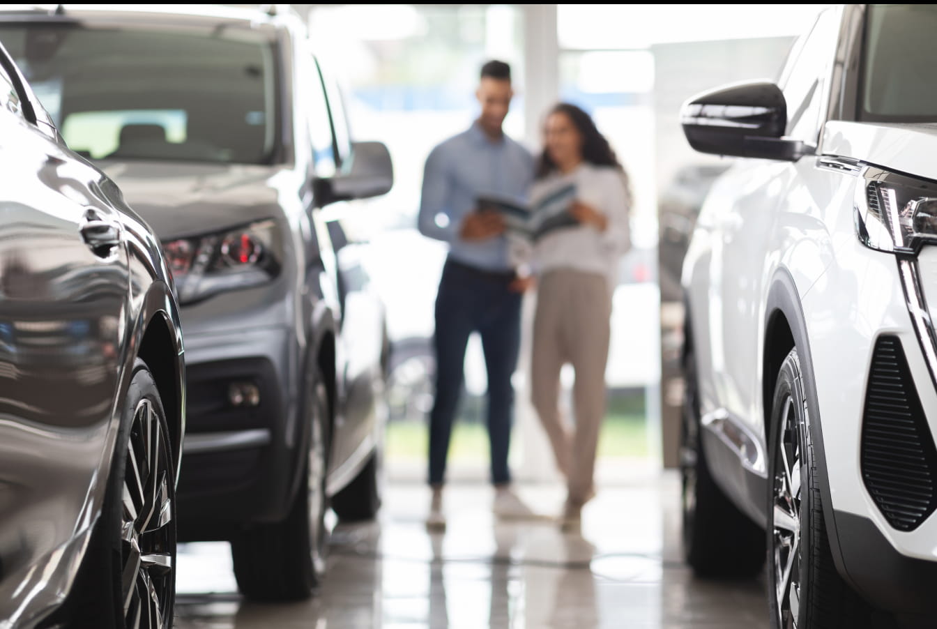 Blurred couple reviews pamphlet in bright car showroom, surrounded by shiny vehicles, conveying excitement and decision-making.