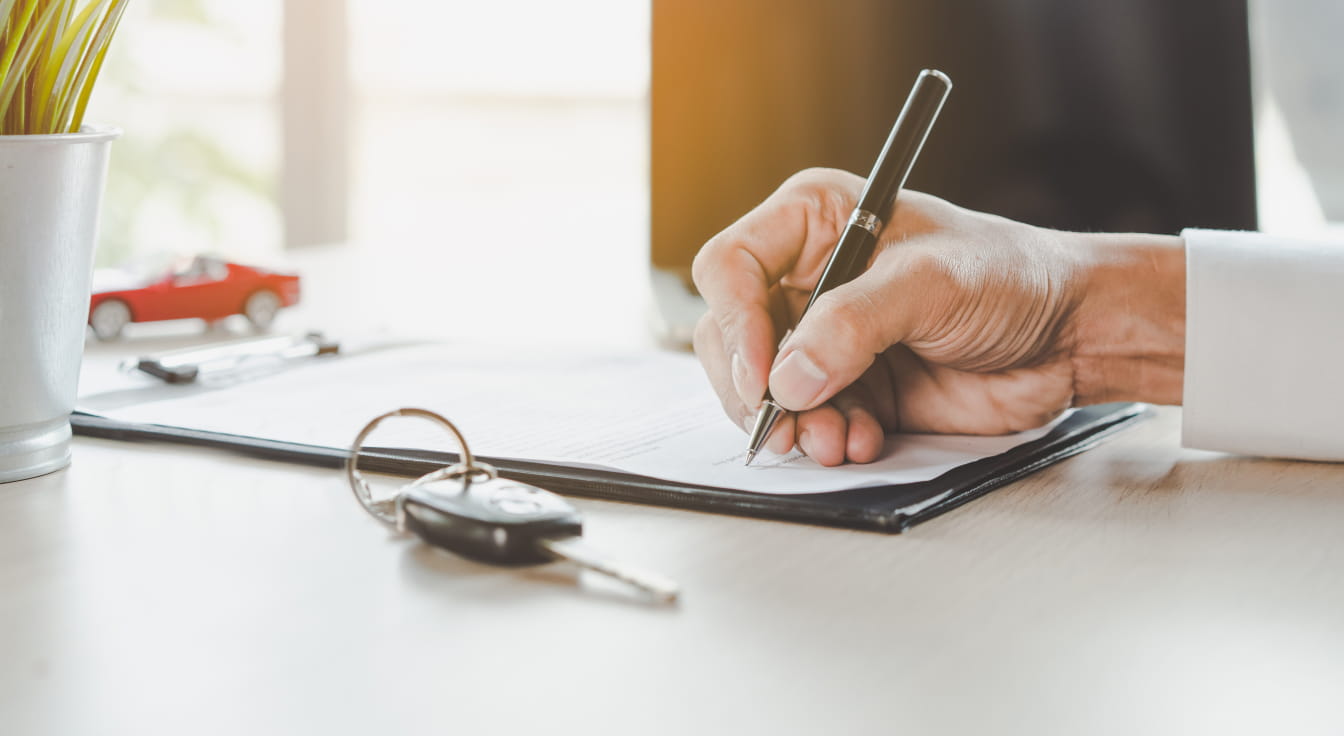 A person signs a document on a wooden desk, with car keys and a small red toy car nearby.