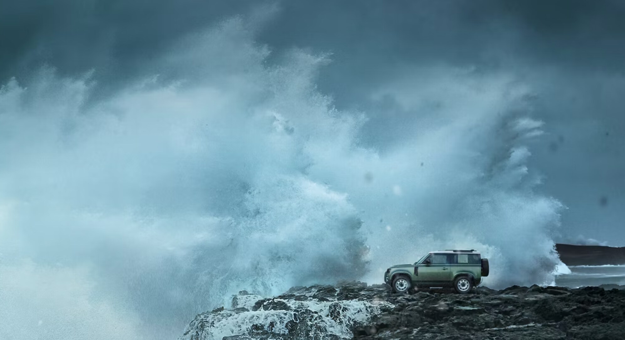 Landscape view of a 2024 Defender parked on a cliff over the ocean with a wave crashing against the edge