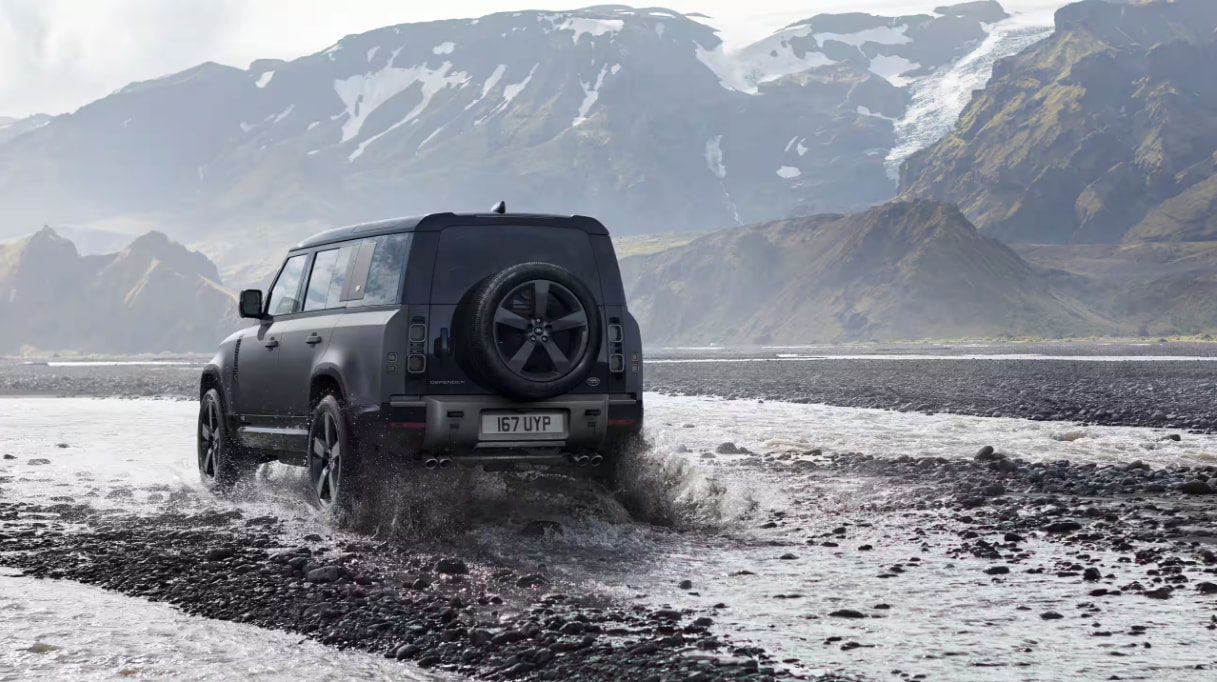 Rear view of the Defender 110 driving through rocks and water with mountains in the background