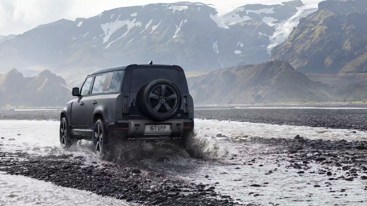 Rear view of the Defender 110 driving through rocks and water with mountains in the background