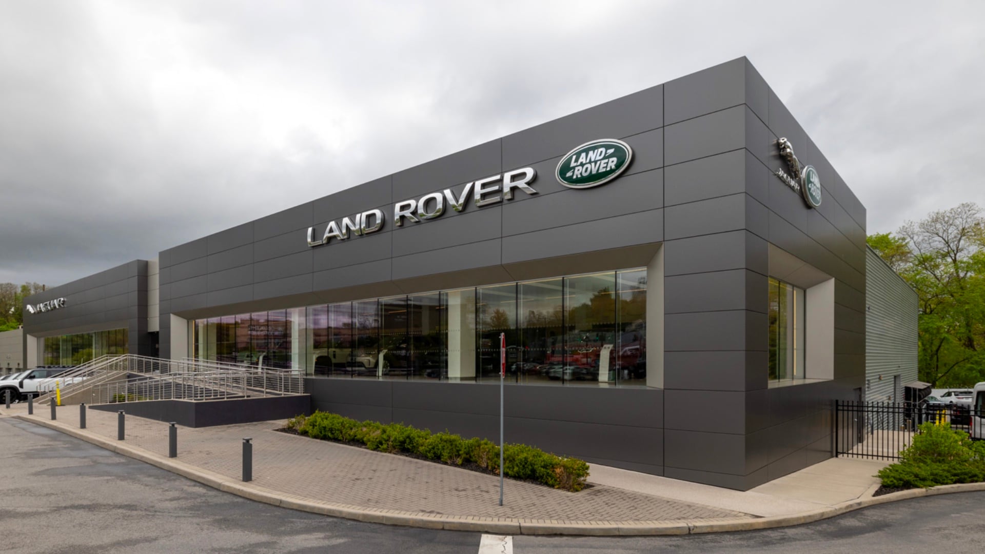 Grey Land Rover White Plains dealership building with a cloudy sky in the background. Vehicles can be seen inside through the large glass windows, and outside in the lot. A large Land Rover logo and sign can be seen on the building.