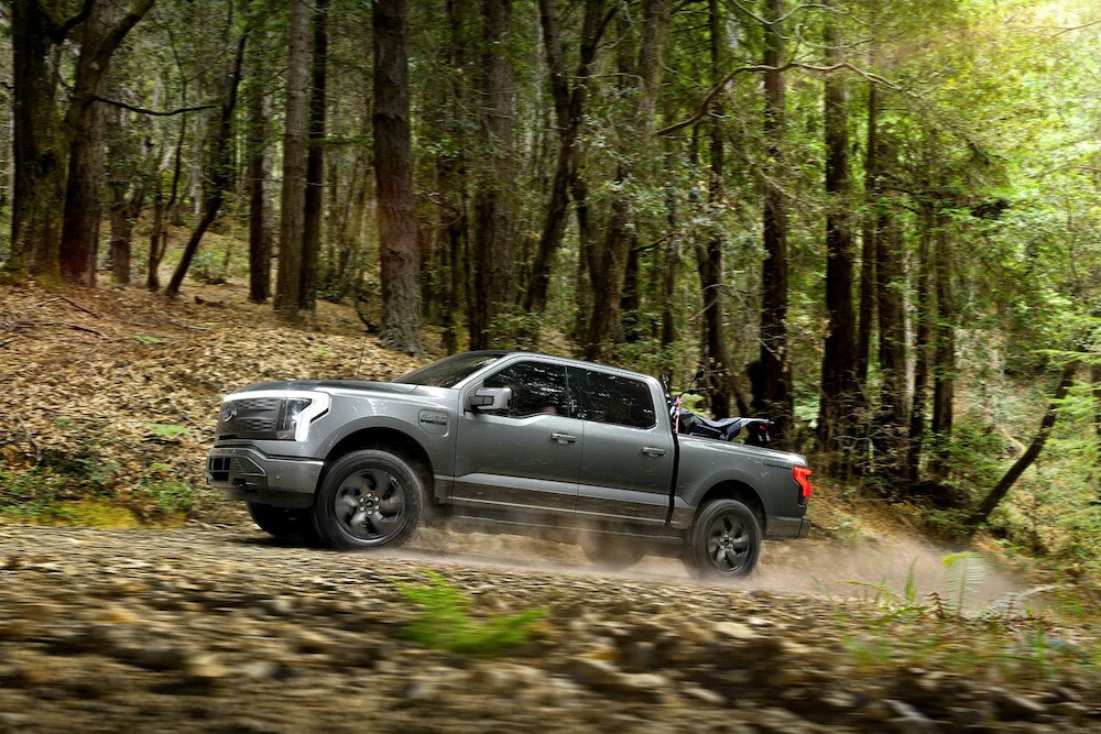 A 2022 Ford® F-150 Lightning being driven along a tree-lined dirt road