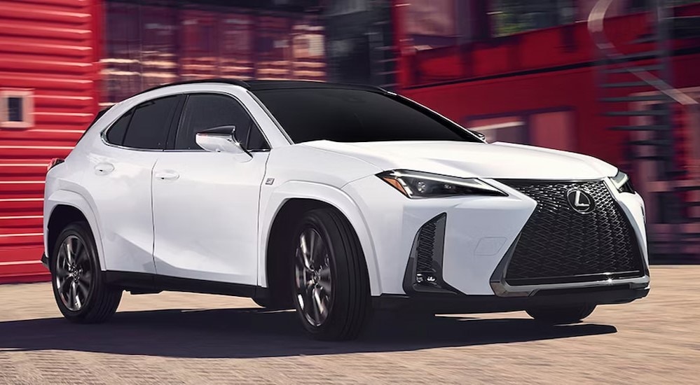 A white 2025 Lexus UX 300h is shown driving near a red building.