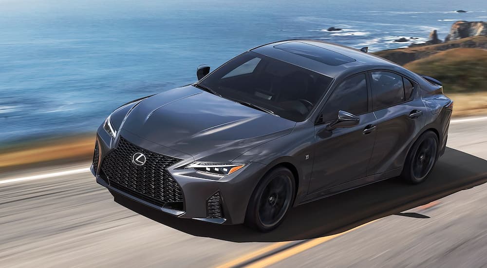 A grey 2024 Lexus IS for sale is shown driving by an ocean.