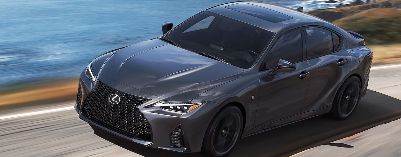 A grey 2024 Lexus IS is shown driving on a road with an ocean view.