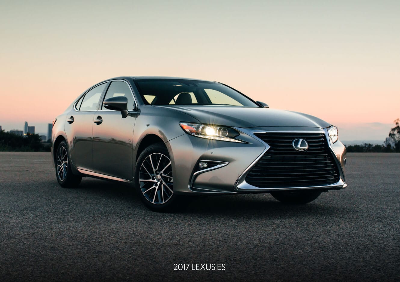 A low exterior front three-quarter view of a silver 2017 Lexus ES parked in a lot against a city skyline at dusk.