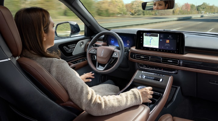 A 2027 Lincoln Aviator® SUV is being driven hands-free on a highway lined with trees. 