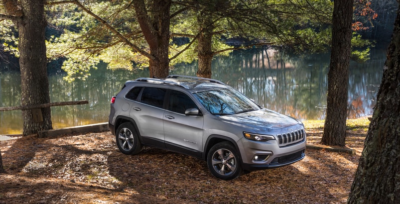 silver Jeep Cherokee SUV parked under forest trees, next to a stream