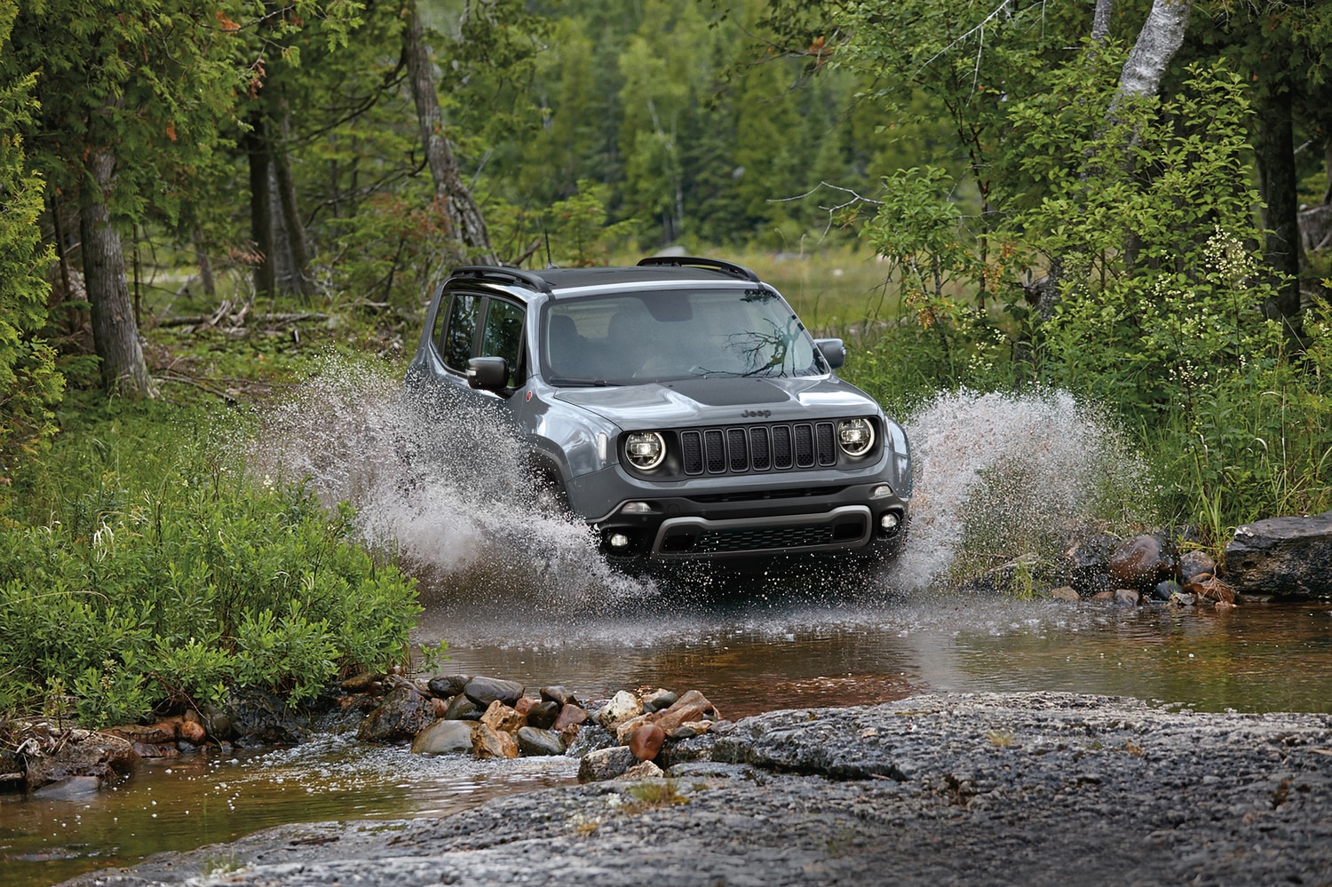 silver Jeep Renegade SUV splashing through a creek in a wooded area
