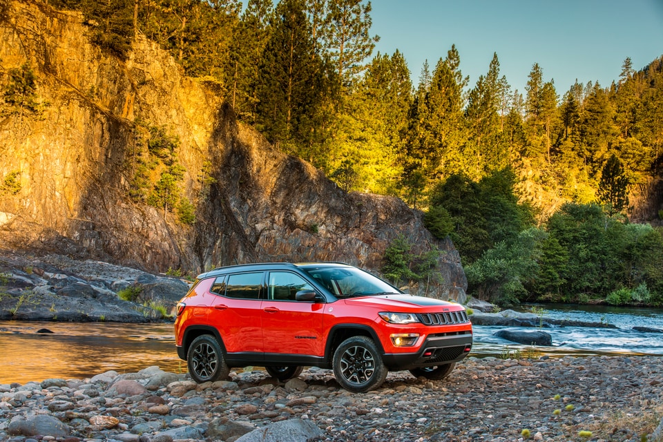 red Jeep Compass SUV parked on a rocky riverside next to a tree-covered mountain