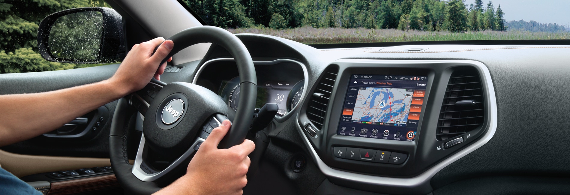 A person holding the steering wheel of a new Jeep Cherokee, the center are of the dash is displaying a map. They are driving in a forest type location.