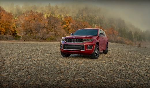 rojo Jeep Grand Cherokee L SUV estacionado en la grava cerca de los árboles de otoño