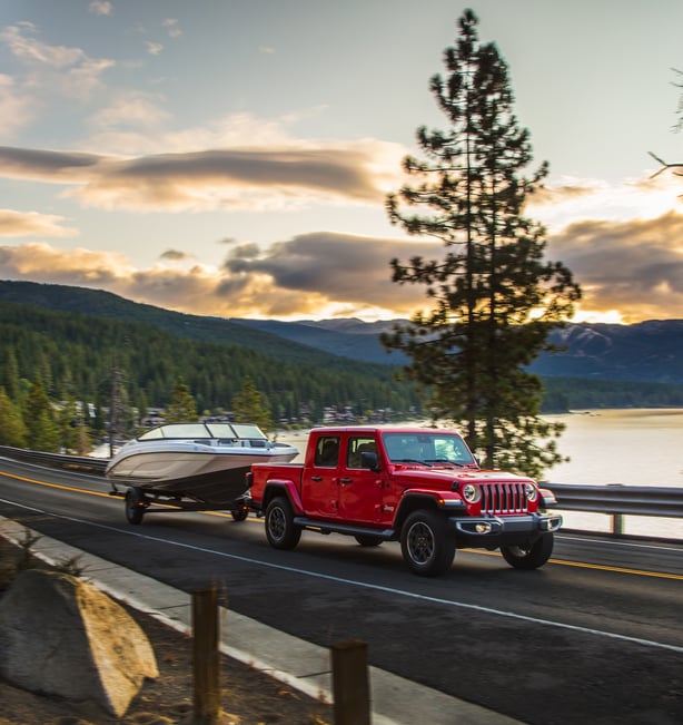 red Jeep Gladiator truck driving down a waterfront highway, towing a boat