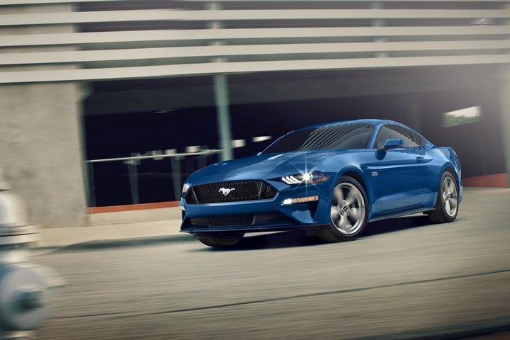 blue Ford Mustang driving fast past a car park