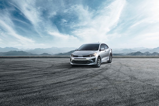 silver Kia Rio sedan parked in an empty lot with a cloudy, blue background