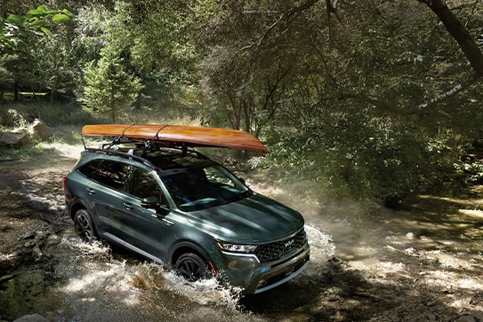 green Kia Sorento driving through a forest creek with a kayak on the roof rack