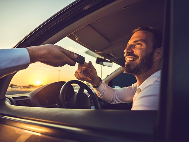 A hand is giving car keys to a smiling person sitting in a vehicle.