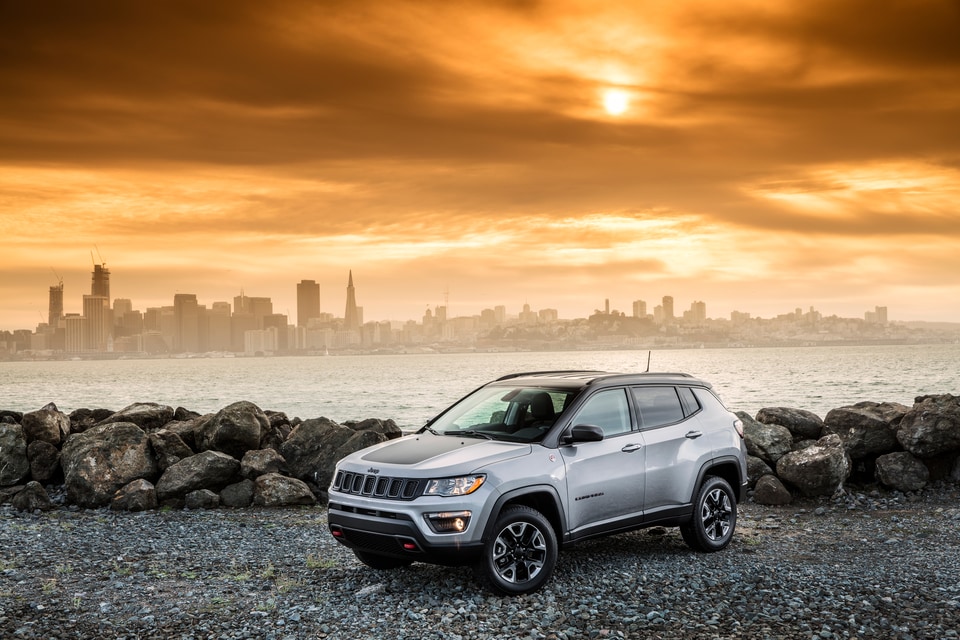 silver Jeep Compass parked next to a rocky shore with a city in the background