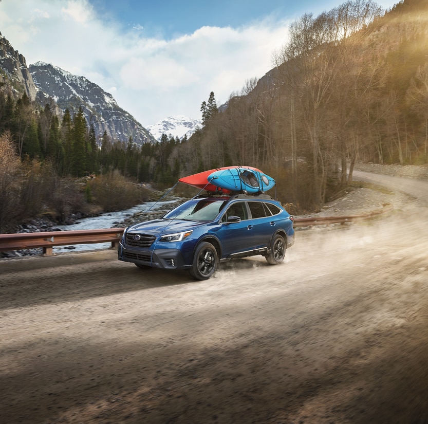 blue Subaru Outback SUV driving on a winding dirt road next to a river, with kayaks on the roof rack