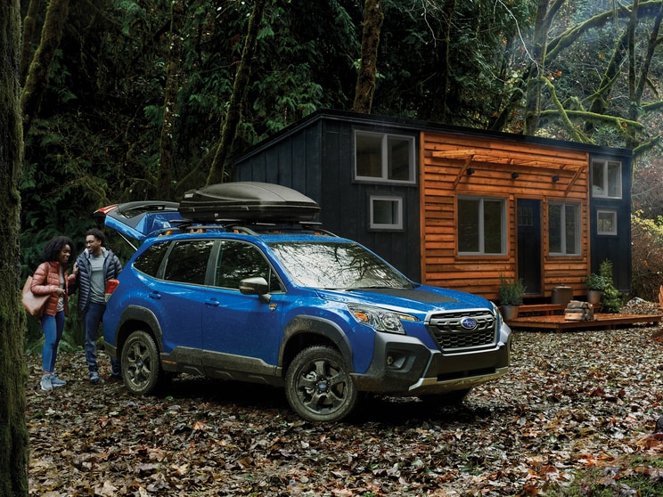 blue Subaru Forester Wilderness edition SUV parked in front of a small cabin with a cargo box on the roof rack