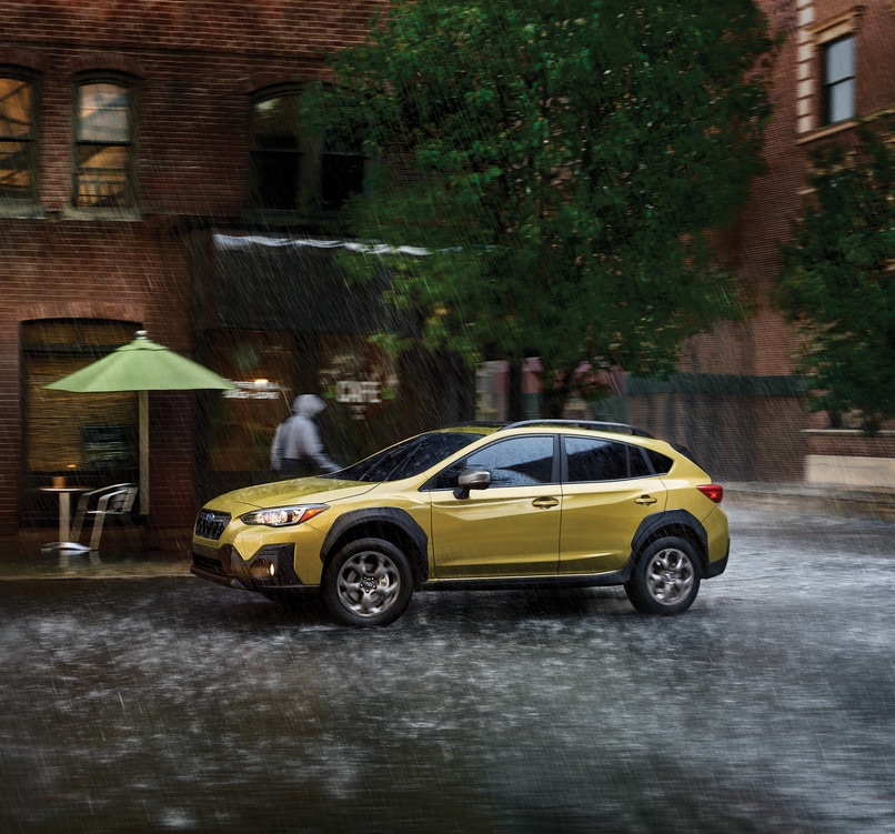 Subaru-Crosstrek-Sport-Hatchback.png yellow Subaru Crosstrek driving past a cafe with an umbrella on a rainy day
