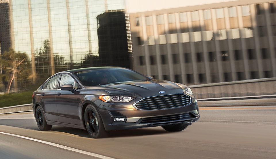 dark gray Ford Fusion sedan driving past a glossy downtown building