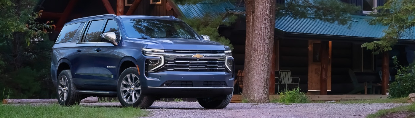 2025 Chevy Suburban 2025 Chevrolet Suburban SUV parked in front of a modern cabin surrounded by large pine trees