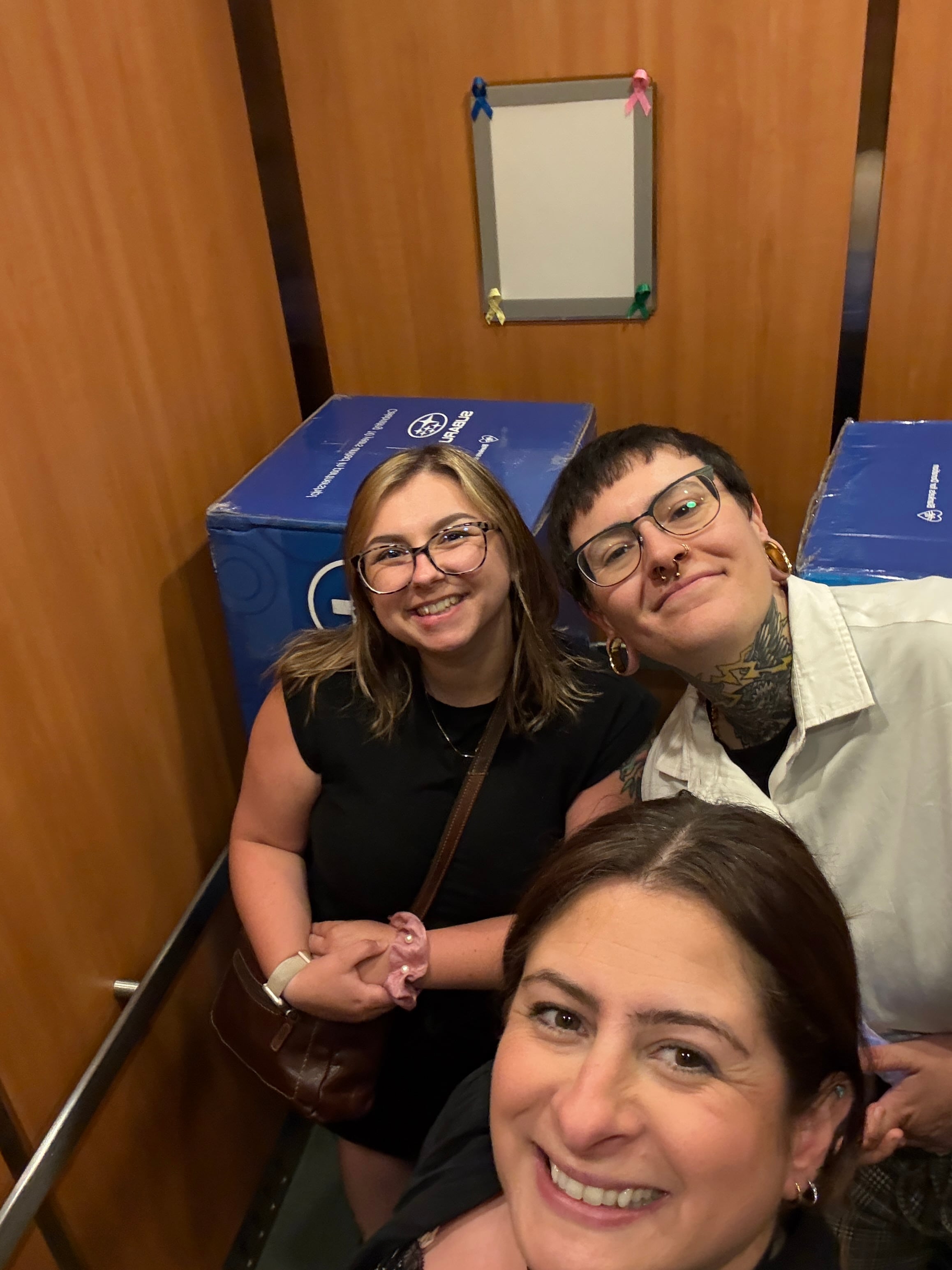 Sam, Carrille and Alexis taking a selfie in the elevator with the blue boxes filled with blankets. 