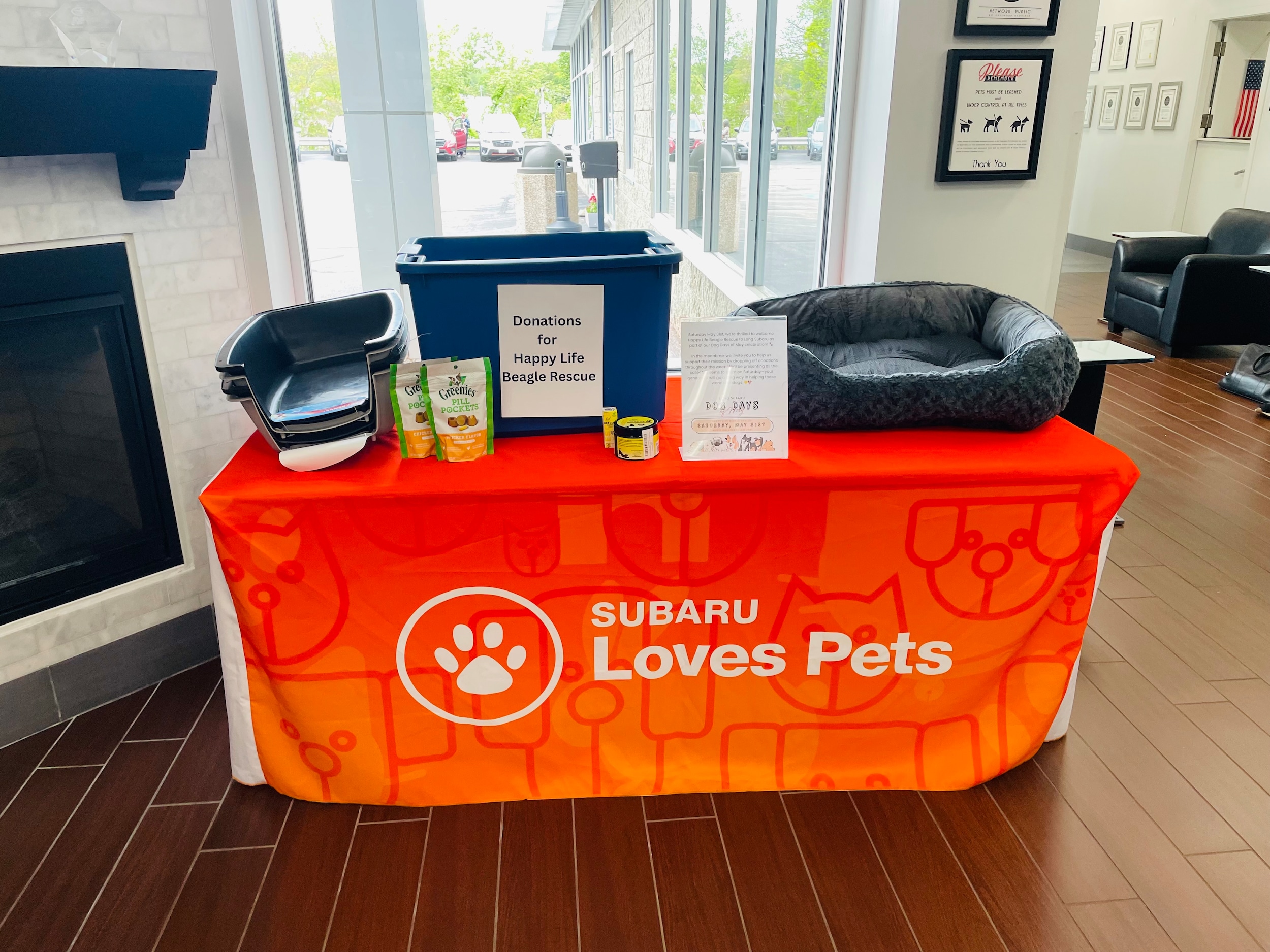 A donation table set up with an orange table cloth saying Subaru Loves pets. there is a navy blue donation bin with items in it. 