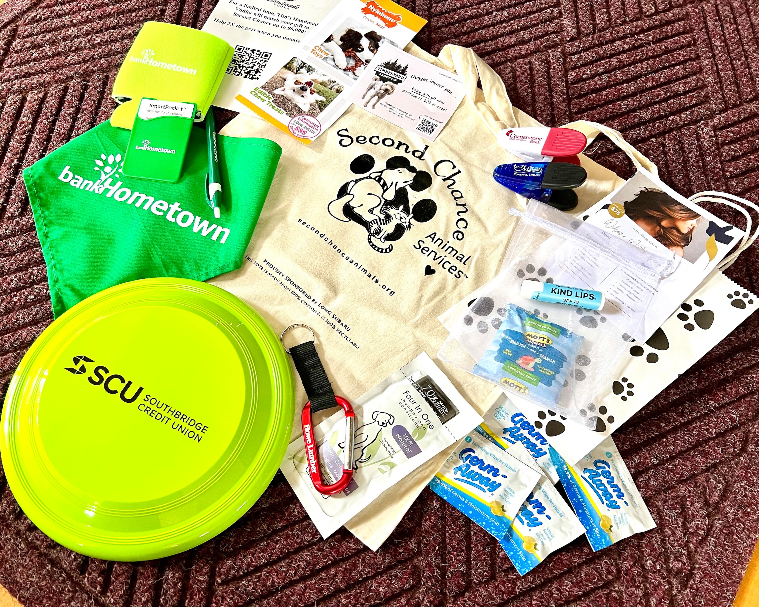 Long Subaru supplies the swag bag as shown in the photo the light tan bag has Second Chance's logo. The bag is filled with a green frisbee, dog bandana and drink koozie. The bag also contains stickers, clips and a keychain.