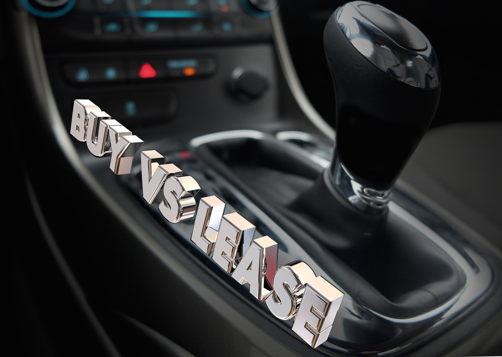 Close-up of a car gear shift with bold 3D text reading “Buy vs Lease” overlaid on the console