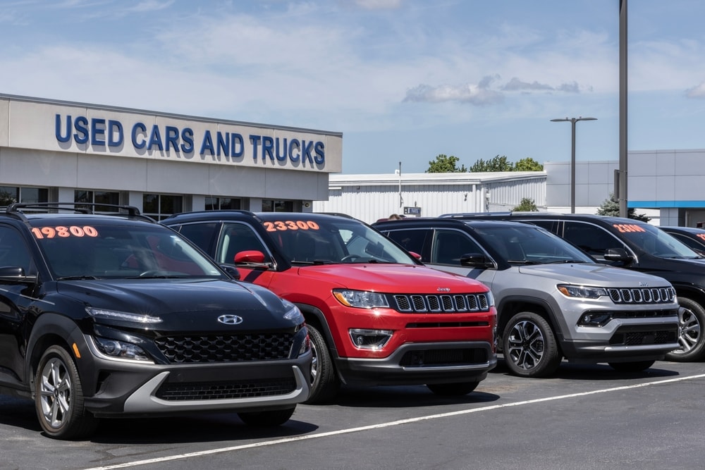 A row of used cars, including black, red, and silver SUVs, parked in front of a dealership with a Used Cars and Trucks sign. Prices are displayed on windshields