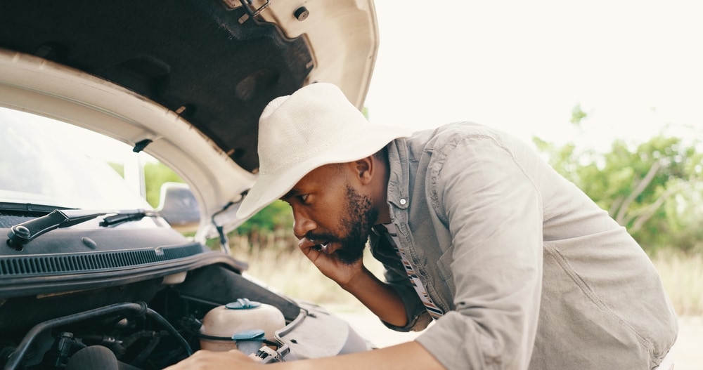 Man wearing a light-colored hat and shirt looks under the hood of a car while talking on his phone, appearing concerned. The car is parked outdoors.jpg