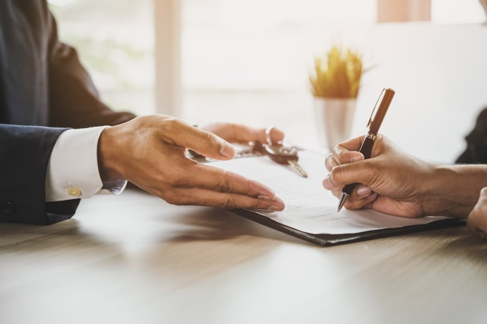The hands of two people, one holding a clipboard of papers, the other signing them