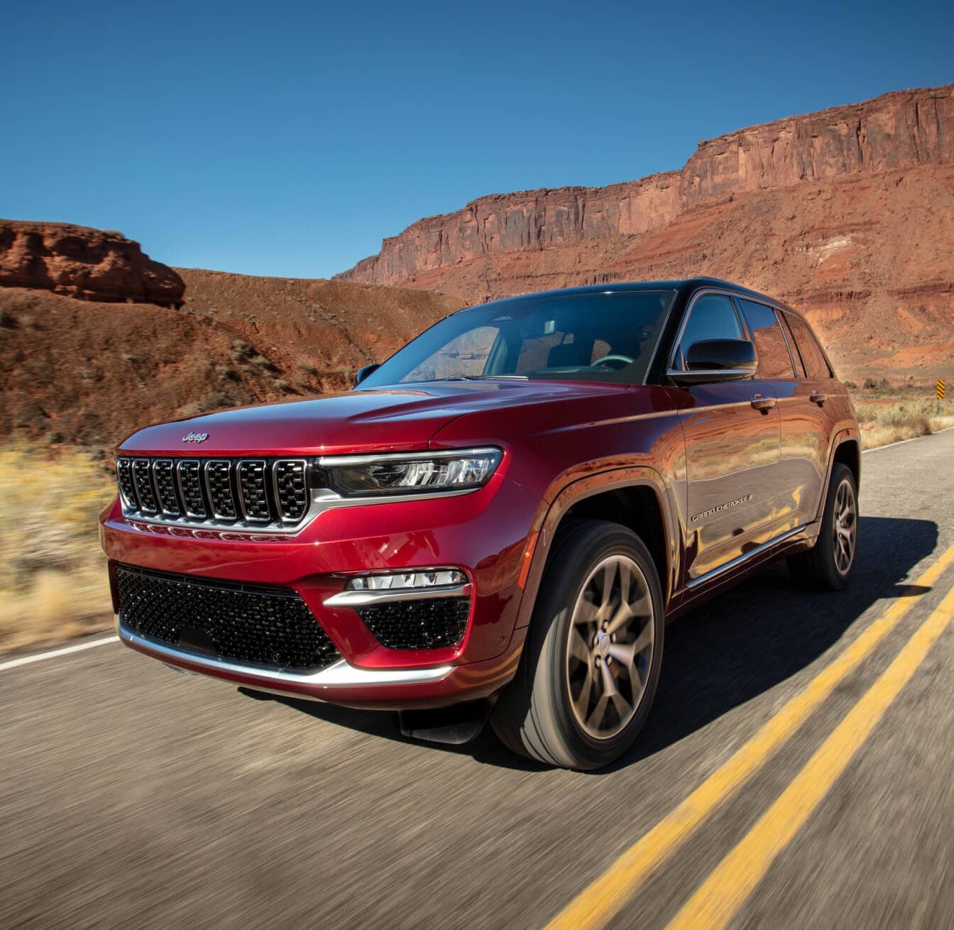 Side three-quarter view of a 2025 Jeep Grand Cherokee in Velvet Red Pearl Driving Through a Desert