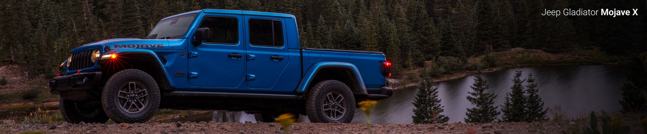 2025 Jeep Gladiator Mojave in Hydro Blue Pearl-Coat off-roading through forest terrain in front of a lake
