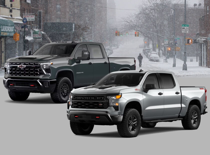Front three-quarter view comparison of a 2026 Chevrolet Silverado 1500 Custom Trail Boss in Sterling Gray Metallic and a 2026 Chevrolet Silverado HD 2500 ZR2 in Cypress Gray against a snowy city.