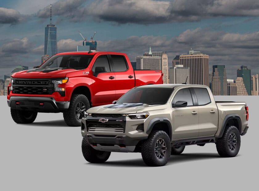 Front three-quarter view comparison of a 2026 Chevrolet Colorado ZR2 shown in White Sands and a Chevrolet Silverado 1500 Custom Trail Boss in Red Hot against a city skyline backdrop.