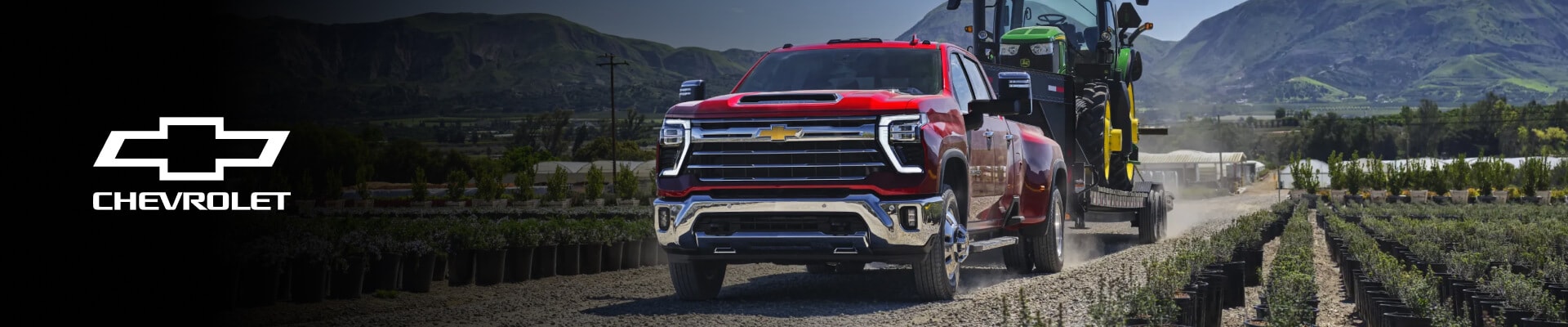 Exterior front three-quarter view of a Red Chevy Silverado HD towing a large tractor down a farmland gravel road.