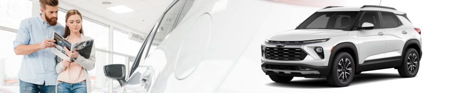 A couple looking over a brochure next to a new car with a white 2026 Chevy Trailblazer superimposed over that vehicle