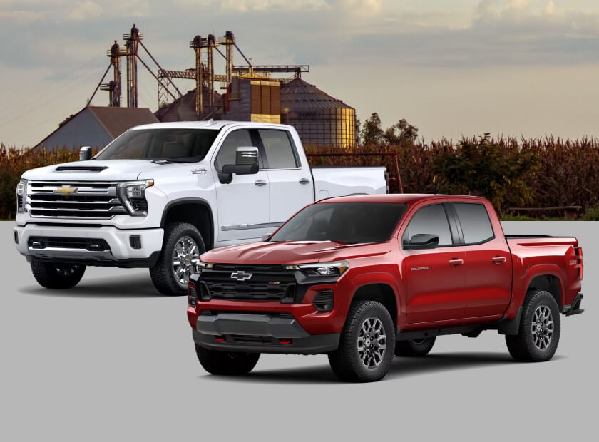 Front three-quarter view comparison of a 2026 Chevrolet Silverado HD 3500 High Country in Polar White Tricoat and a 2026 Chevrolet Colorado Z71 in Radiant Red Tintcoat against a farm skyline.